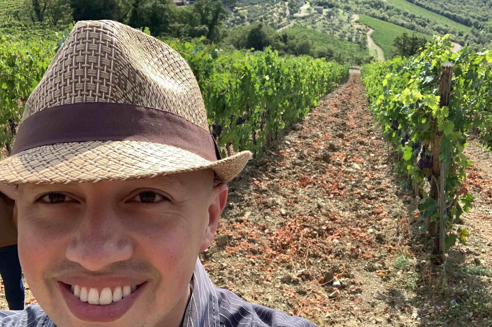 Alex in a straw fedora standing between rows of Sangiovese vines in a Chianti Classico vineyard, with Tuscan hills and a stone village in the background.